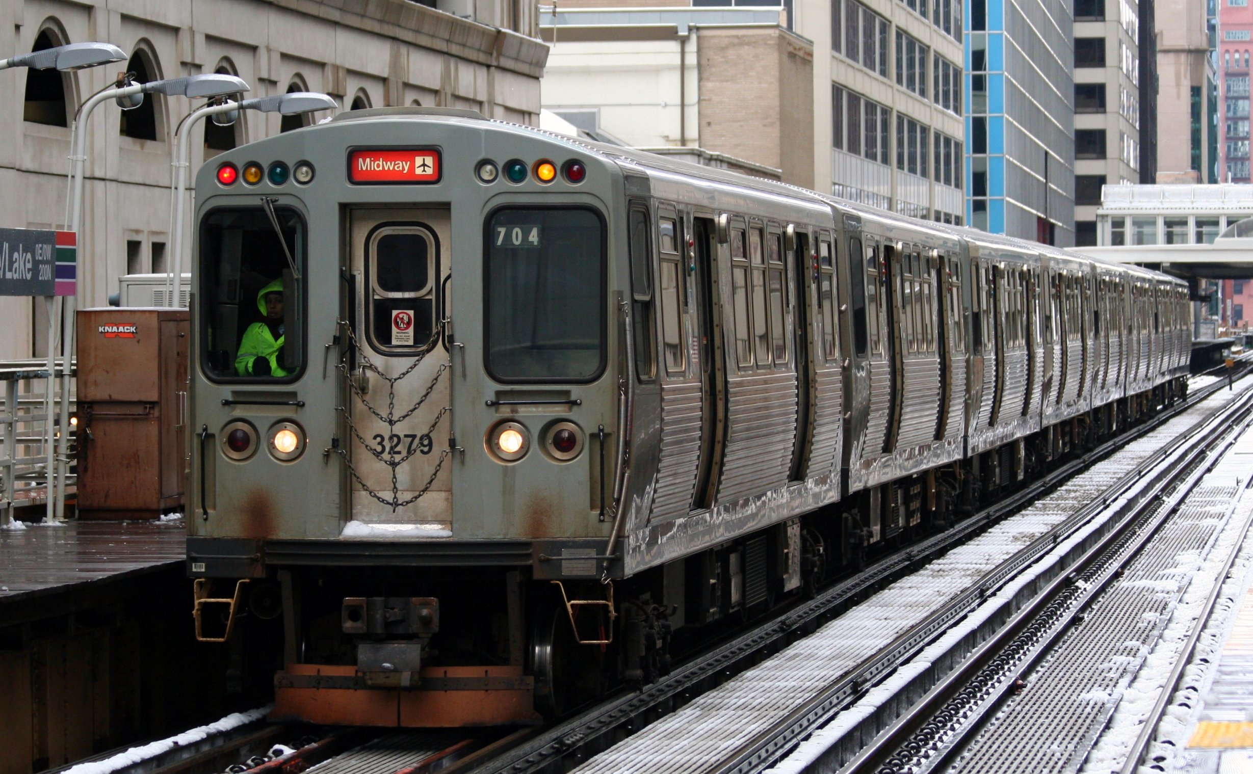 A Chicago Transit Authority (CTA) Orange Line train bound for Midway Airport stops at the elevated State/Lake station in downtown Chicago. The silver railcars have a slightly weathered exterior, and a train operator in a bright green jacket is visible in the cab. Snow dusts the tracks and platform, and tall city buildings surround the scene.